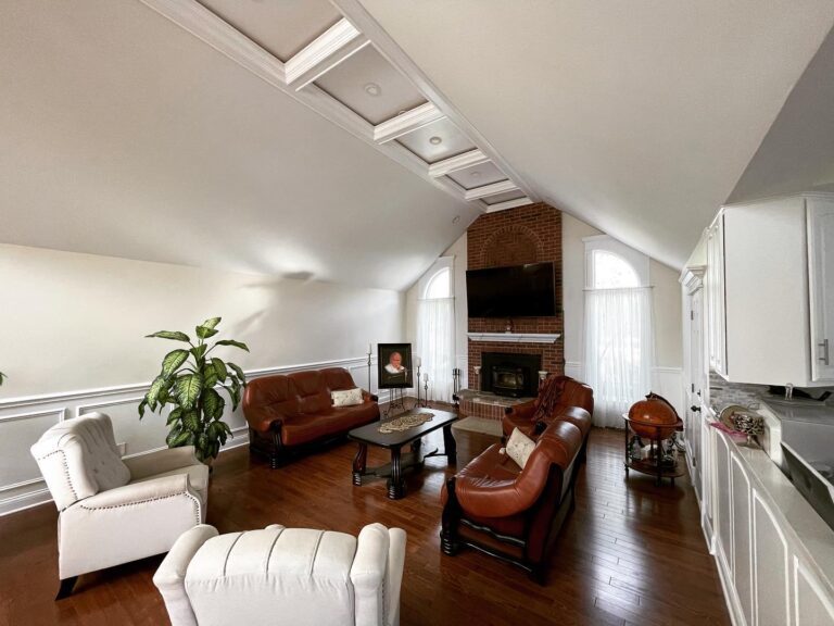 Stylish and inviting living room featuring a comfortable gray sectional sofa, a mid-century modern coffee table, a plush area rug, a wall-mounted flat-screen TV, and large windows with sheer curtains, accented by indoor plants and contemporary wall art.