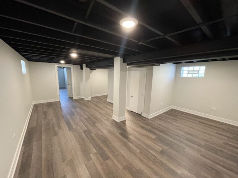 A refreshed, empty basement renovation space with new flooring panels after comprehensive basement renovation services. The walls are painted in a neutral color, adding brightness and spaciousness to the interior. Ceiling lights illuminate the room, highlighting its modern appeal and showcasing the quality of renovation work.